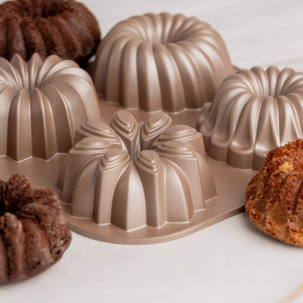 A set of six non-stick bundt cake pans in a metallic taupe color, shown with several baked chocolate and fruit cakes on a light marble surface.