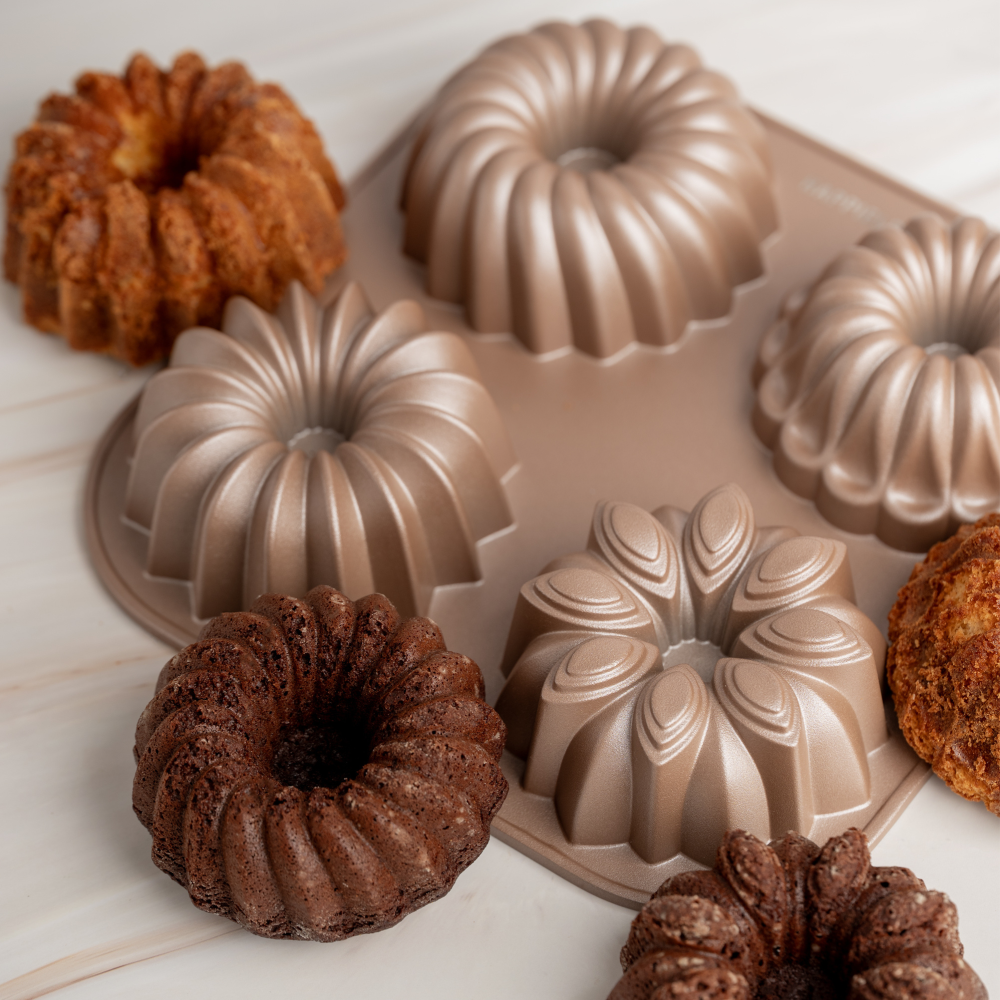 A silicone bundt cake mold set shown with several baked ring-shaped cakes on a light marble surface. The set includes a rose gold non-stick mold with multiple fluted and floral-shaped cavities, alongside three finished brown cakes used as examples.
