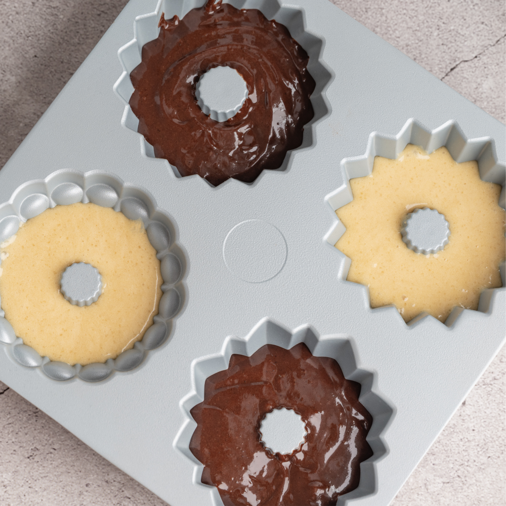 A square gray silicone baking mold with four scalloped ring-shaped cavities, shown partially filled with baked goods: two golden-brown ring cookies and two chocolate-topped ring cookies, on a light stone surface.